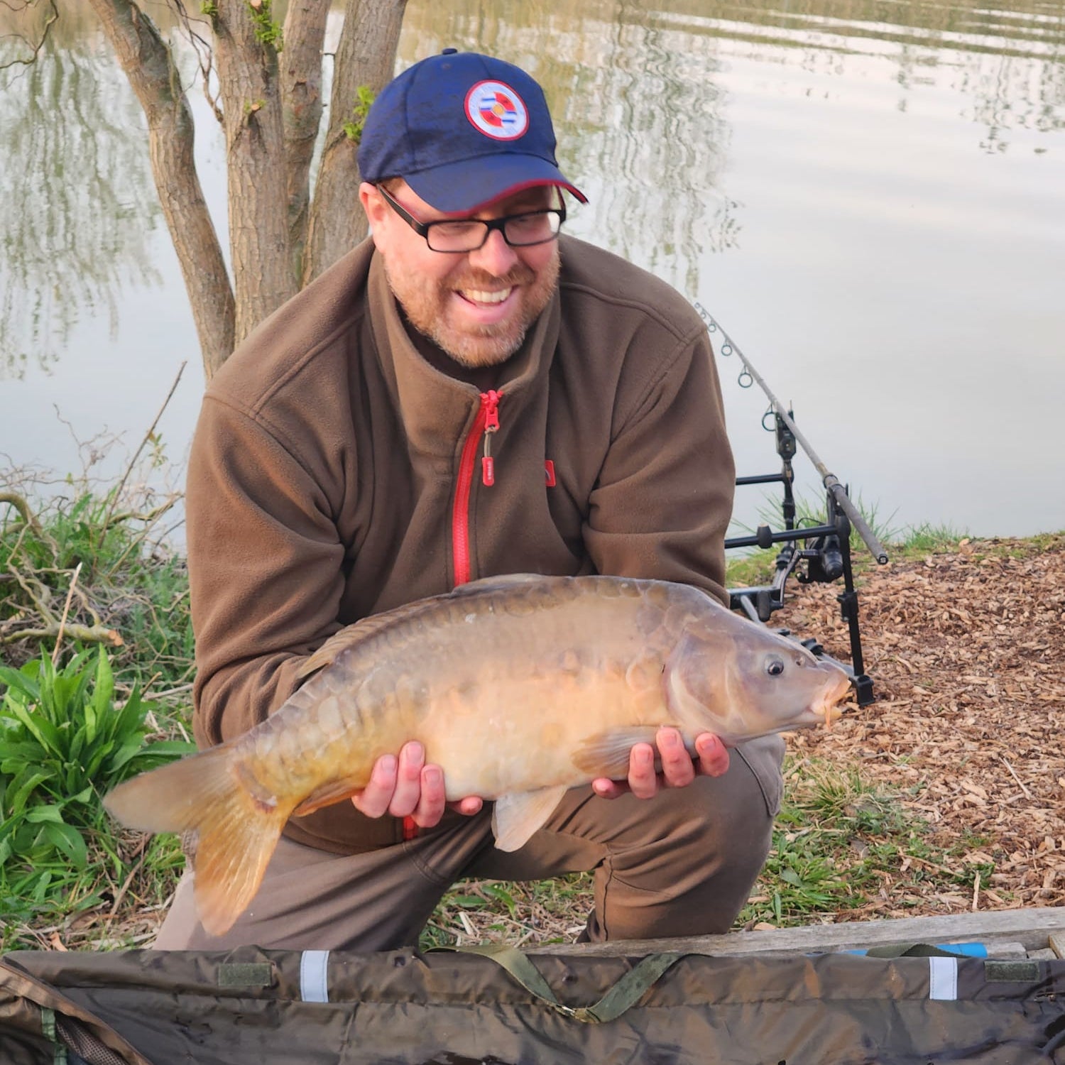 Man holding a large fish by a lake with fishing equipment in the background