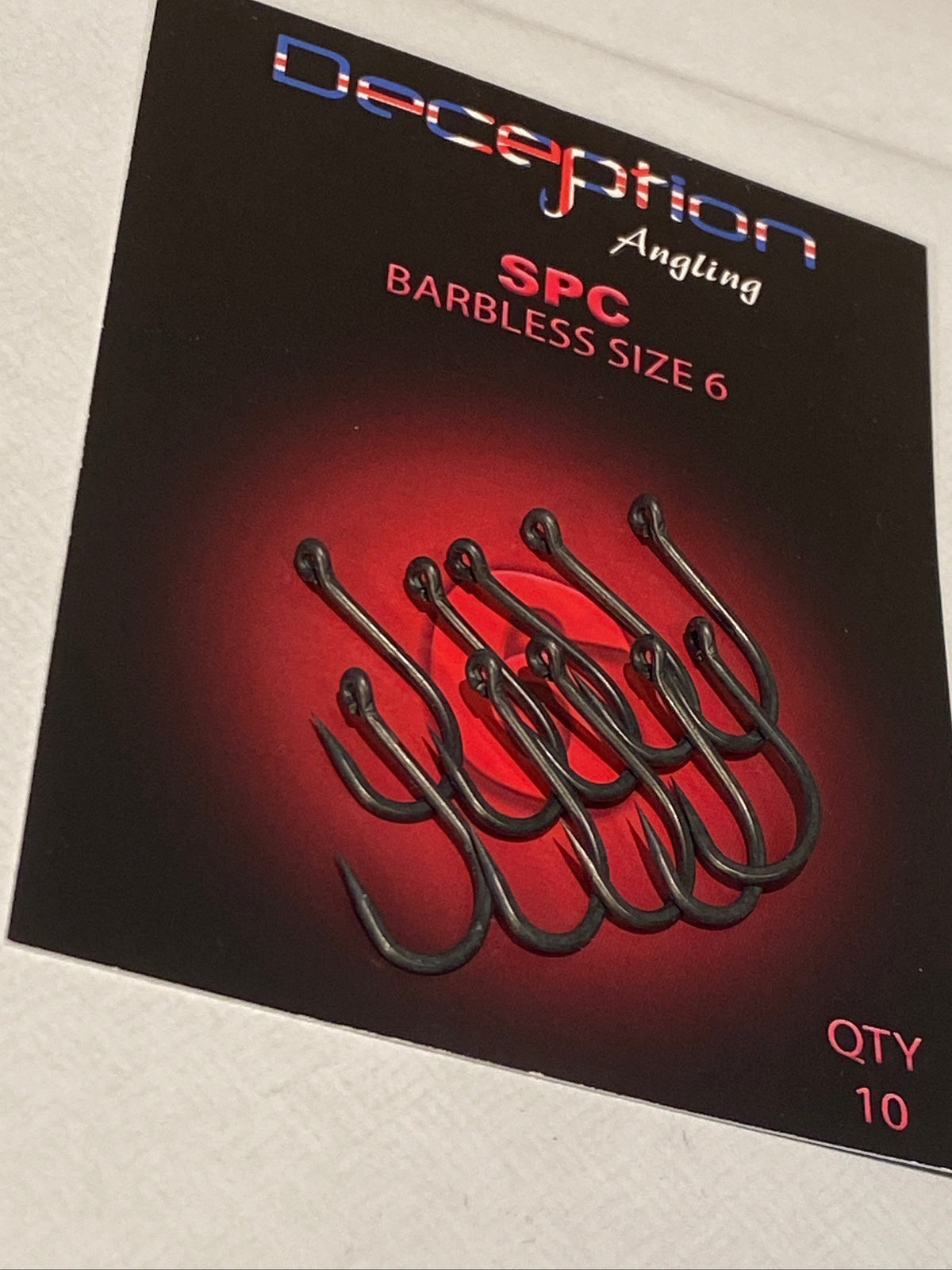 A package of Deception Angling SPC Barbless Size 6 hooks, featuring a black card with a red glow and an illustration of six dark hooks, placed on a white textured surface.